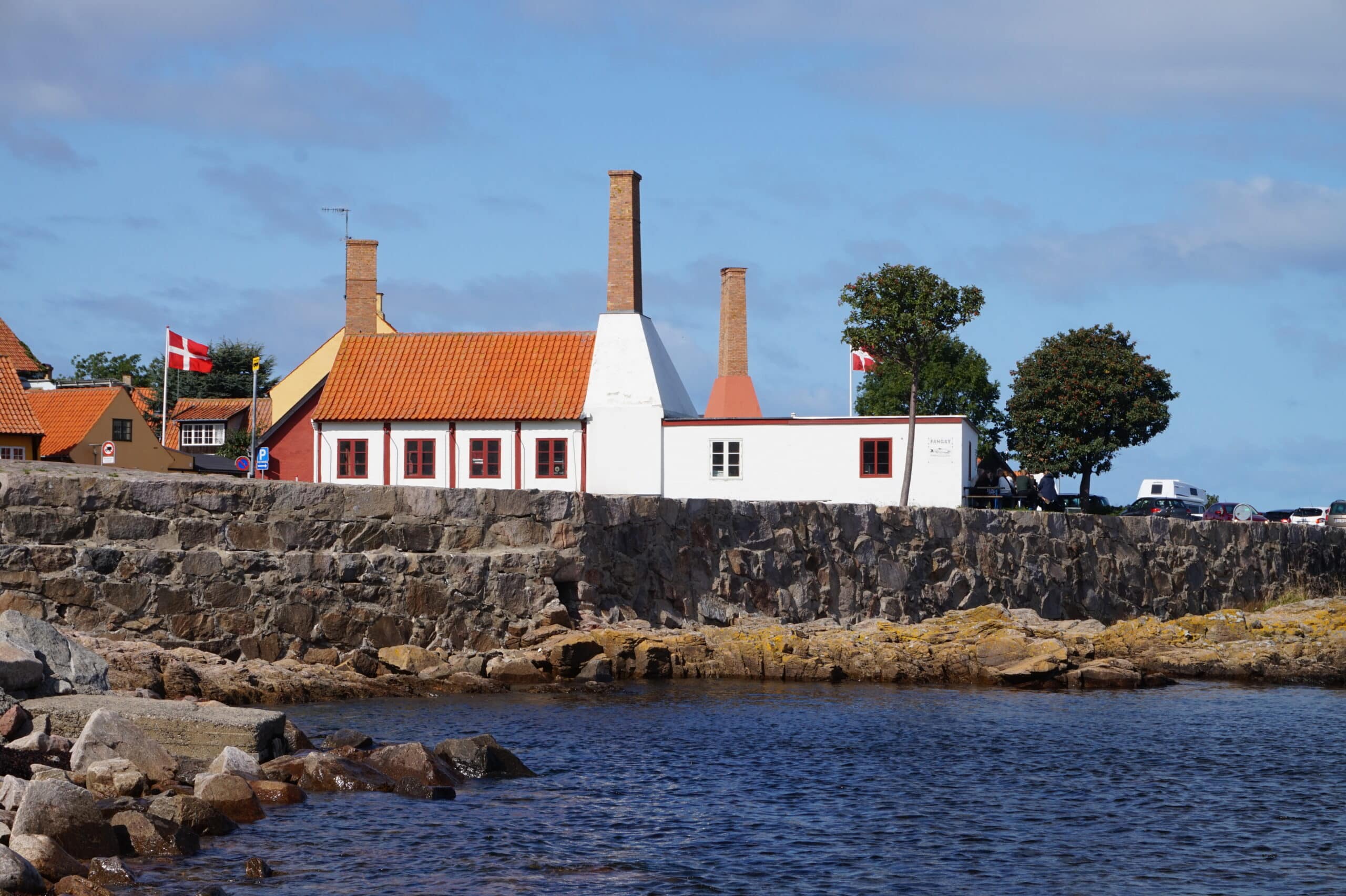Weiße Räucherei mit orangefarbenem Dach, Backsteinschornsteinen und dänischer Flagge hinter einer Steinmauer am Meer