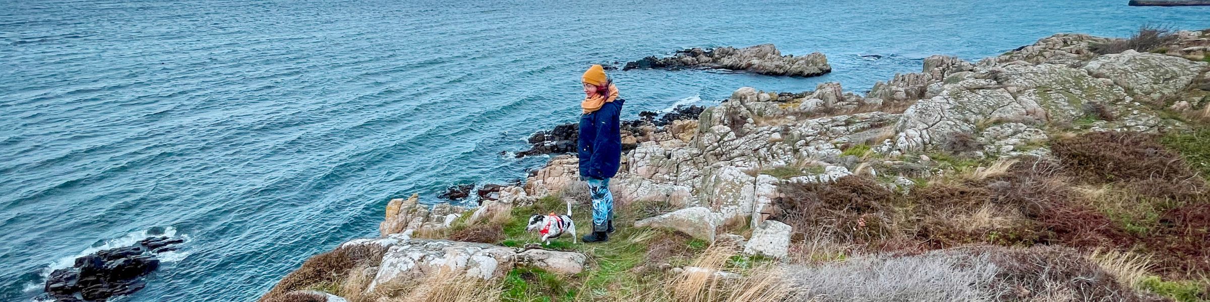 Ewelina in orangefarbener Mütze und Schal mit einem kleinen Hund im roten Geschirr auf Felsen am Meer
