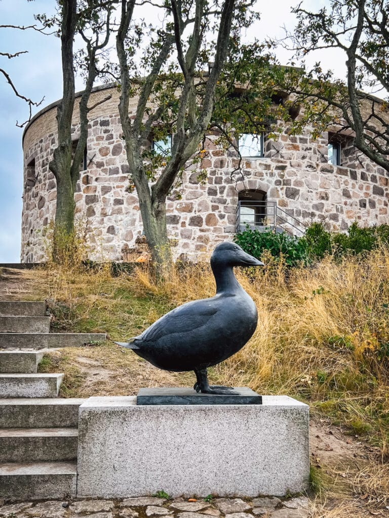 Skulptur einer Eiderente auf einem Sockel vor einem runden Steinturm