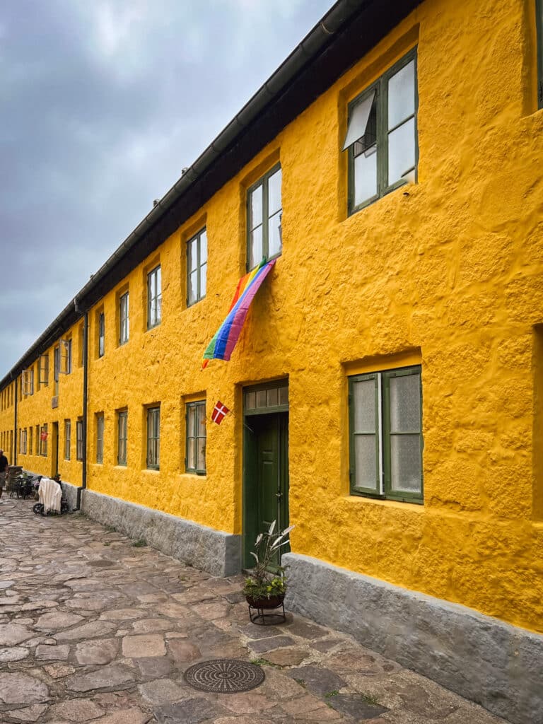 Langes gelbes Gebäude mit grünen Fenstern und Regenbogenfahne an einer gepflasterten Straße