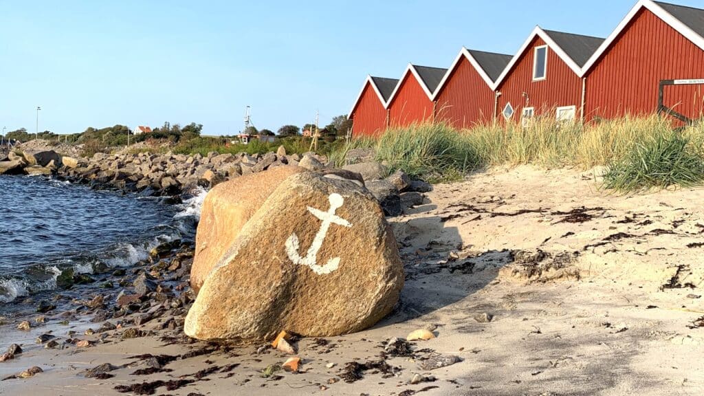 Felsblock mit aufgemaltem Anker am Strand vor roten Holzhäusern