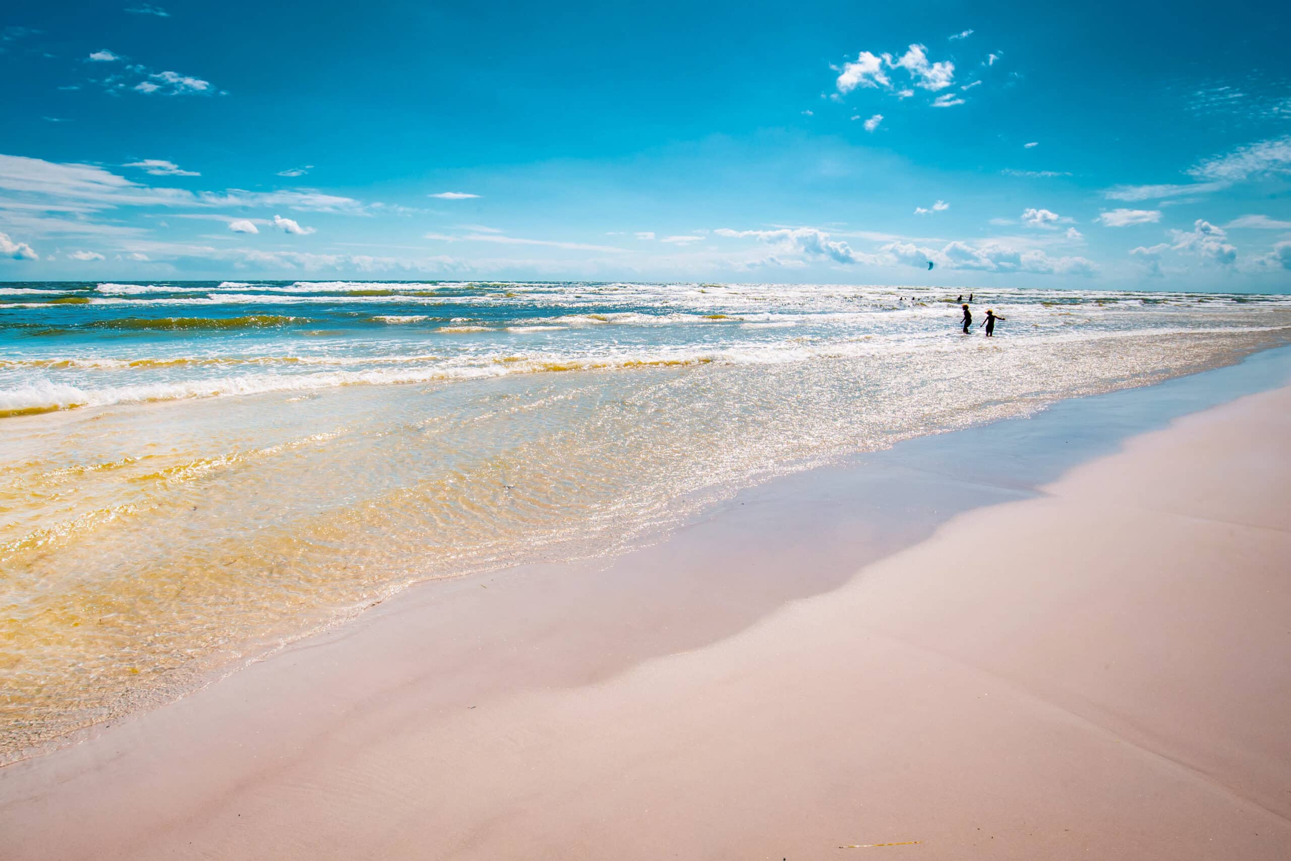 Breiter Sandstrand mit flachem, klarem Wasser, in der Ferne zwei Personen in den Wellen und Kites von Kitesurfern