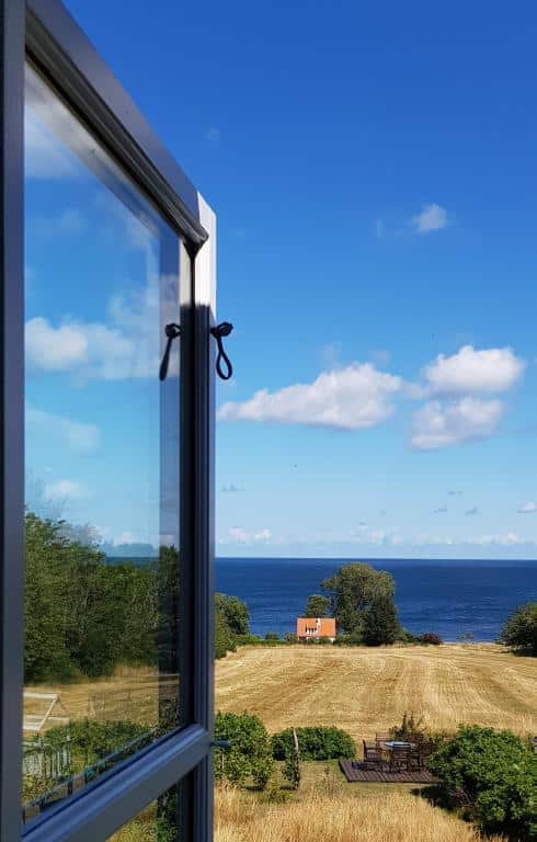 Blick aus einem offenen Fenster auf ein Feld, ein Häuschen mit rotem Dach und die Ostsee