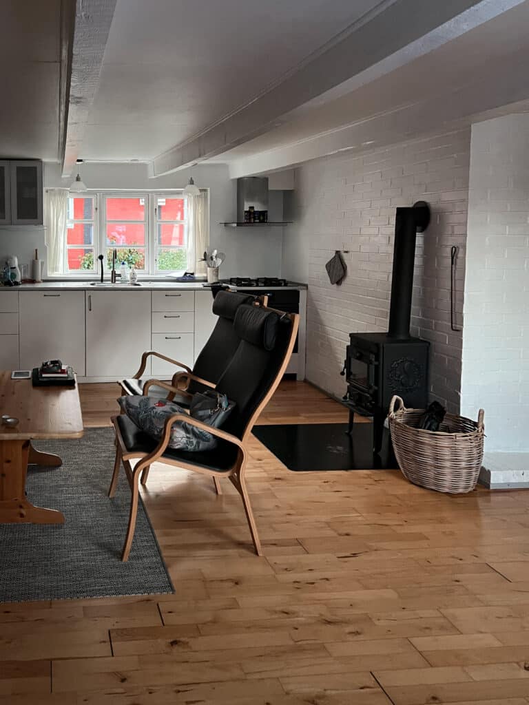 Heller Wohnraum mit Küchenzeile, Deckenbalken, Sessel und schwarzem Holzofen