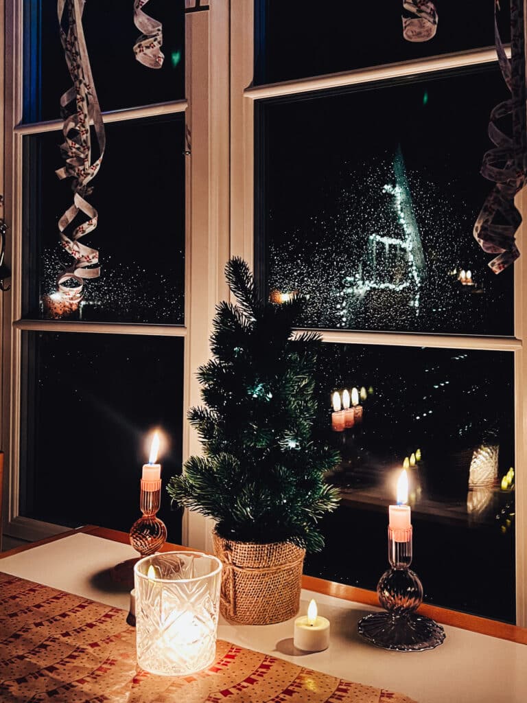 Kleiner Weihnachtsbaum, Kerzen und Luftschlangen am Fenster, hinter der nassen Scheibe ein beleuchteter Giebel