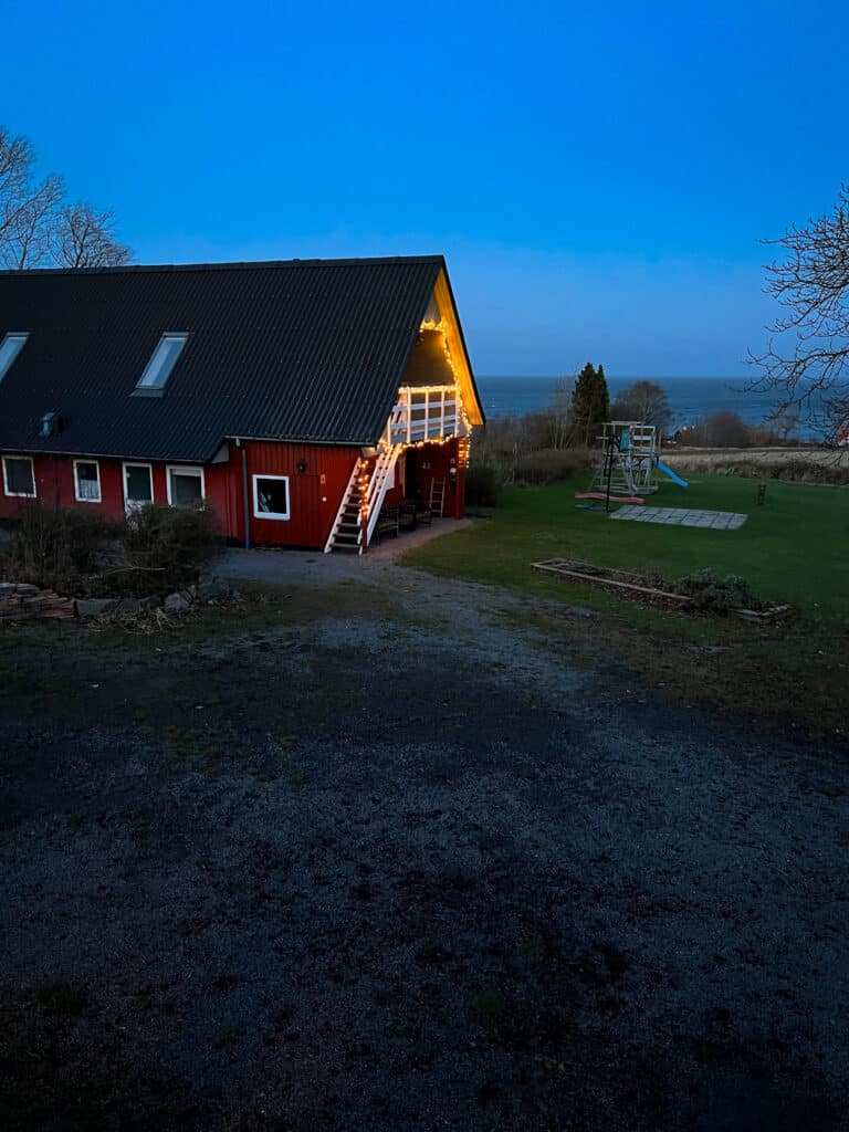 Rotes Gebäude mit Ferienwohnungen in der Dämmerung, am Giebel eine Weihnachtslichterkette, daneben ein Spielplatz, im Hintergrund die Ostsee