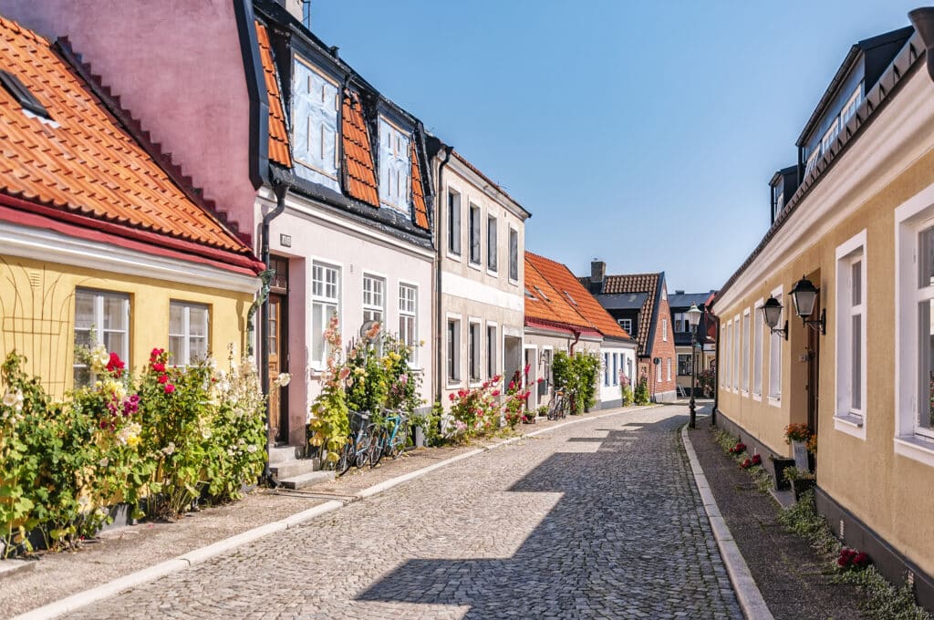 Gepflasterte Gasse in Ystad mit bunten Häusern, orangefarbenen Dachziegeln und Stockrosen an den Wänden
