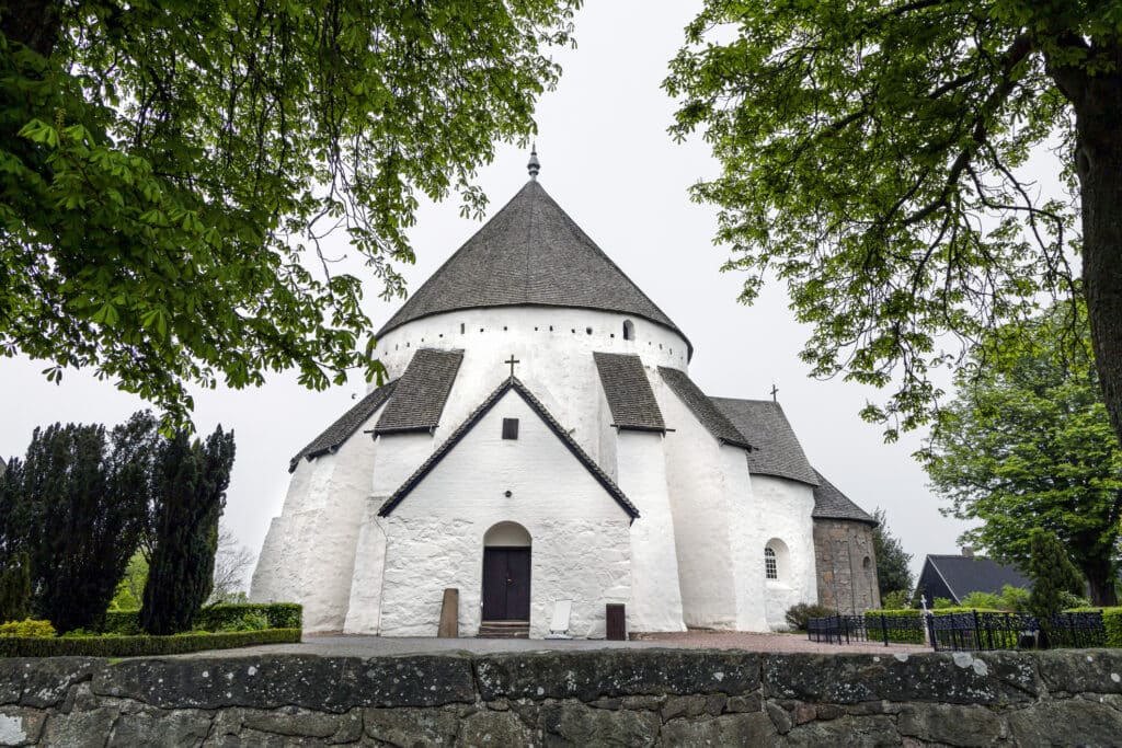 Weiße Rundkirche in Østerlars mit Kegeldach und Stützpfeilern, dahinter eine Steinmauer und Bäume
