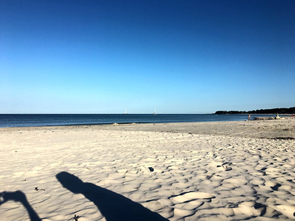 Weißer, welliger Strandsand in der Abendsonne mit langen Schatten zweier Personen, dahinter die Ostsee mit Segelbooten