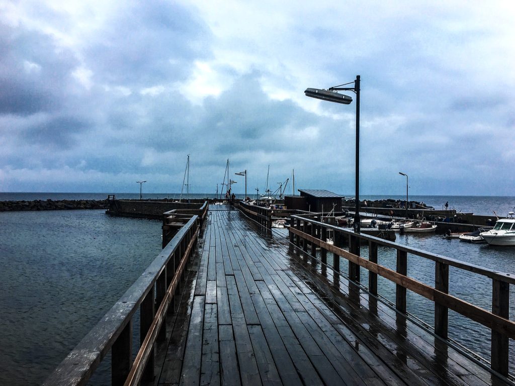 Nasser Holzsteg mit Geländer führt zu einem kleinen Hafen mit Booten, darüber Regenwolken
