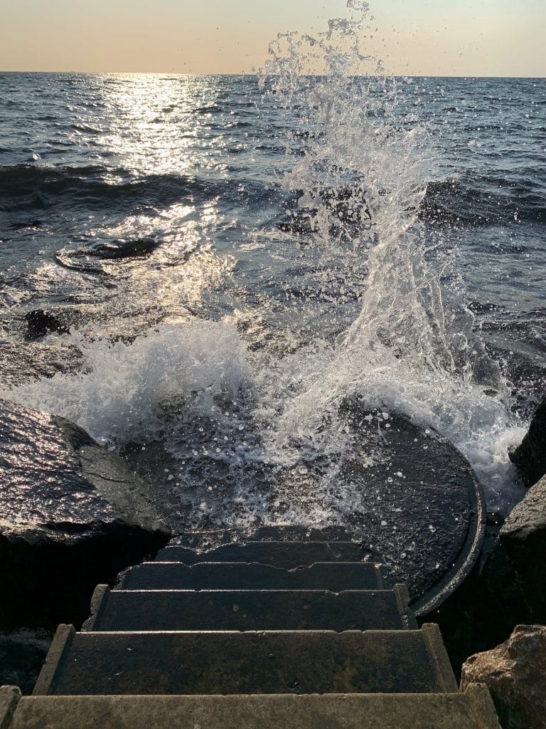 Eine Welle bricht sich an der Treppe ins Meer in Hasle, Nachmittagssonne über dem Wasser