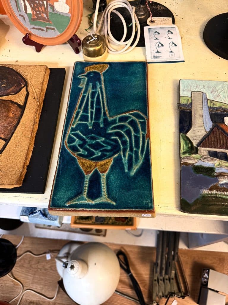 Keramikfliese mit blauem Hahn, Bilder und Lampen auf einem Tisch im Laden Kunst Kuriosa