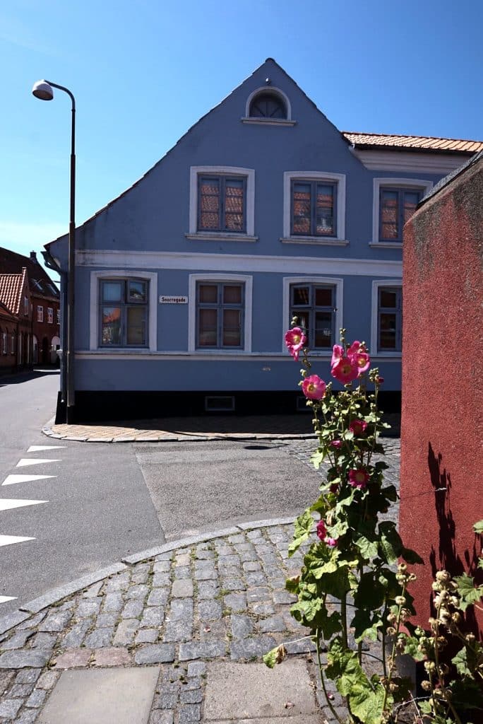 Blaugraues Eckhaus in Rønne, davor blühende Stockrosen und Kopfsteinpflaster