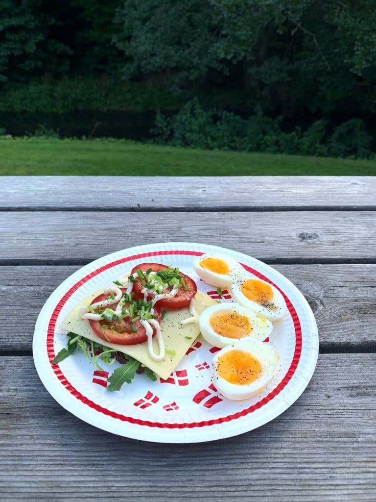 Sandwich mit Käse, Tomate und Rucola und Eier auf einem Pappteller mit Dänemark-Flaggen, auf einem Holztisch mit Blick auf Rasen und Bach