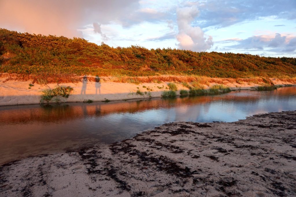 Düne im goldenen Abendlicht spiegelt sich im Bach, auf dem Sand die Schatten von zwei Personen
