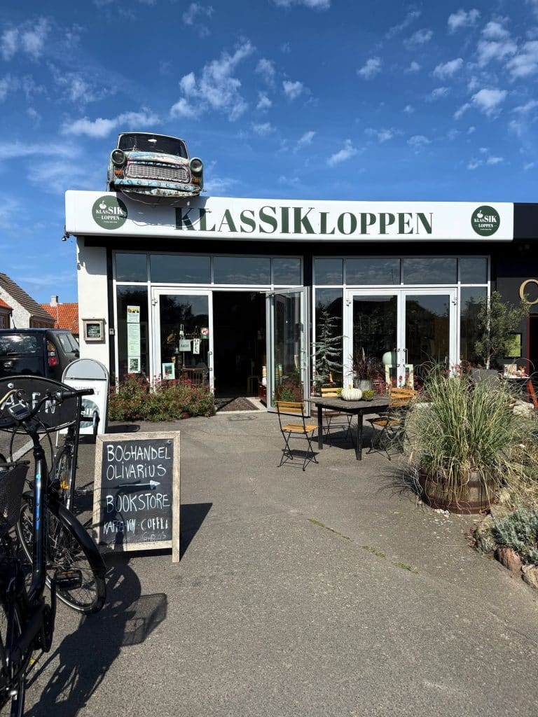Weißes Gebäude mit dem Flohmarkt Klassikloppen in Svaneke, auf dem Dach ein altes Auto, vor dem Eingang Tische und Fahrräder